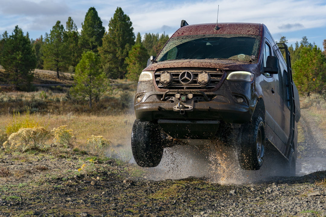 Front jounce reinforcement bracket for Sprinter AWD (2023-present) displayed with a car on a dirt road, highlighting rugged off-road capability and essential suspension support.