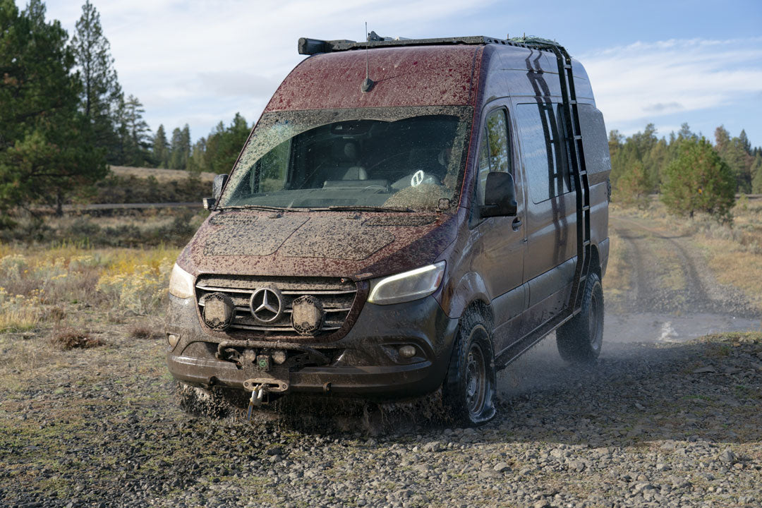 Front jounce reinforcement bracket for Sprinter AWD (2023-Present) shown installed on a van navigating a muddy terrain, highlighting durability and support in rough conditions.