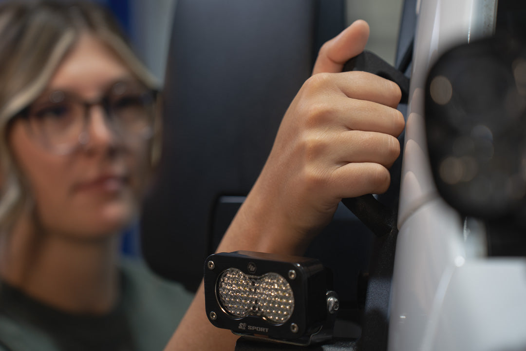 Person wearing glasses gripping an A-Pillar Grab Handle Light Mount on the side of a white 2019+ Sprinter Van, with a compact LED light mounted below their hand in focus.