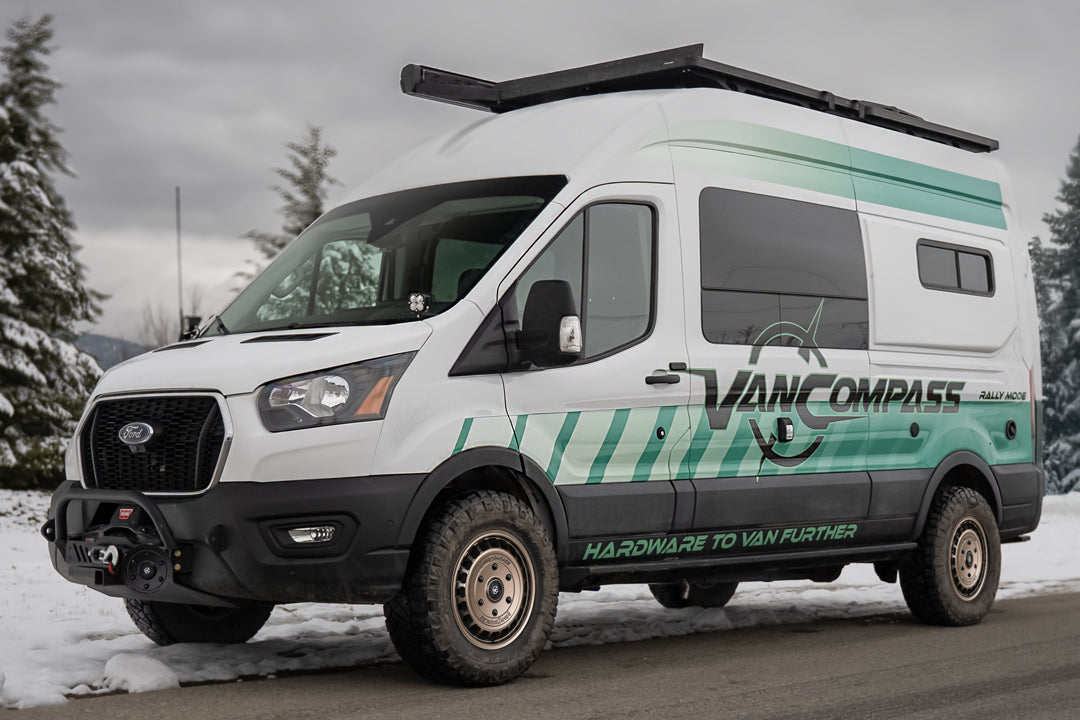 A white Ford Transit camper van with teal and black graphics, a metallic bronze-colored wheel rim with a multi-spoke design and cutouts, featuring the 'Van Compass' brand name and logo embossed on the center cap, parked outdoors on a snowy road with trees in the background.
