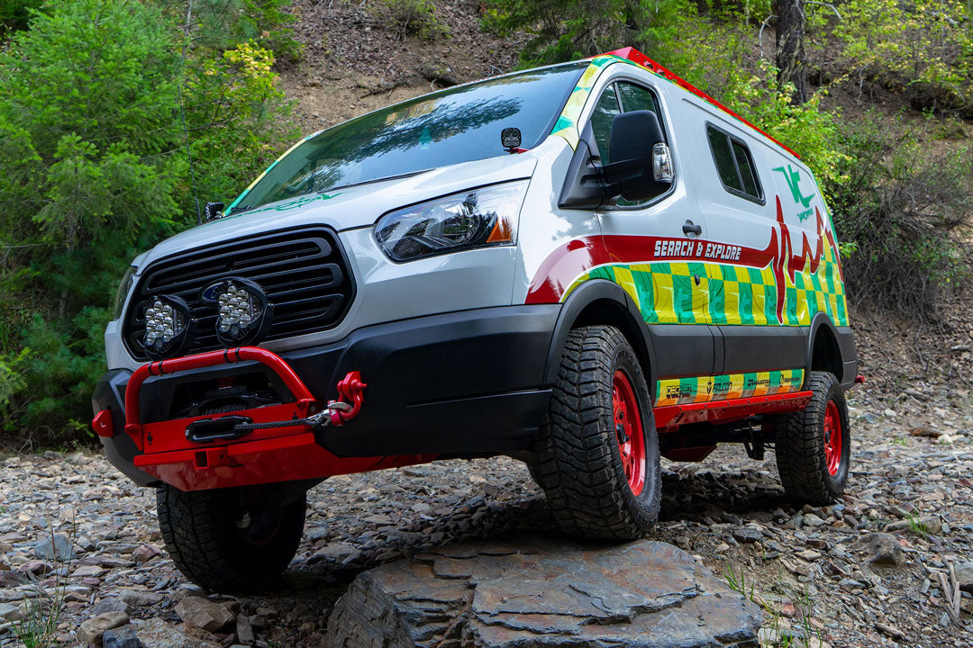 Van equipped with VAN COMPASS COMPLETE 4 LIFT KIT - TRANSIT (2015-PRESENT RWD SINGLE WHEEL ONLY), shown navigating a rocky road, highlighting enhanced ground clearance and larger tires.