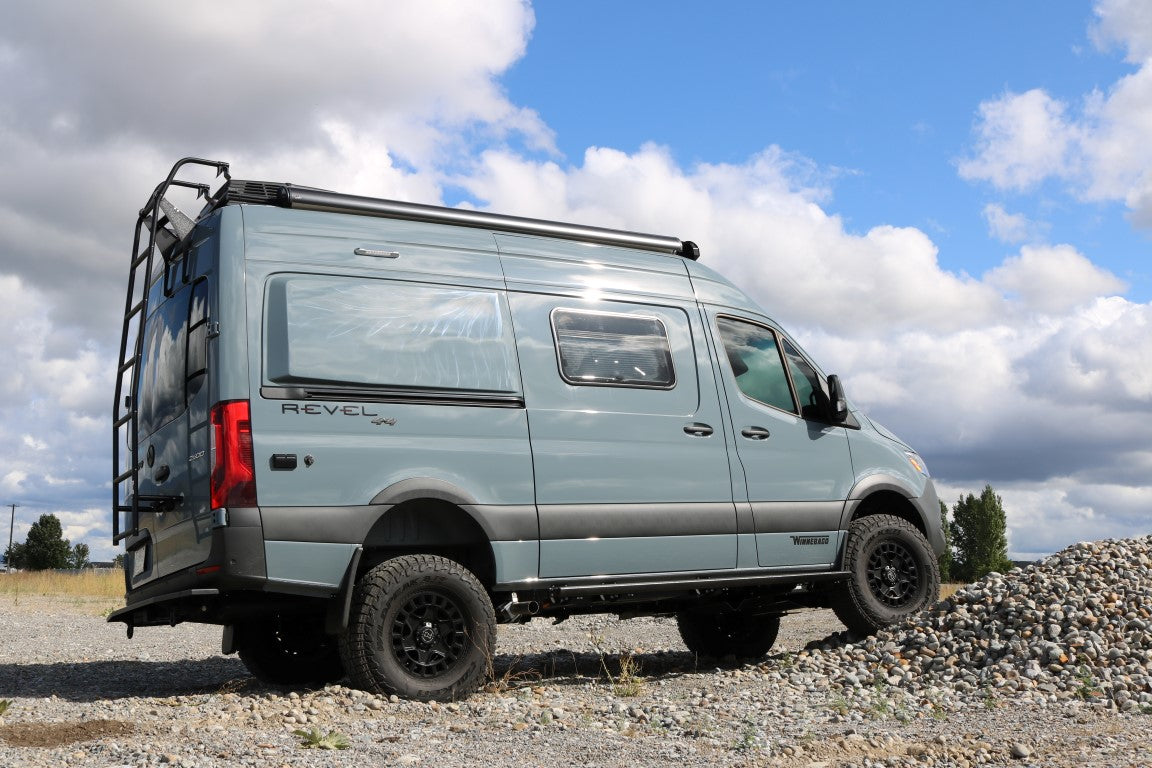 BIG TIRE MUDFLAP RELOCATION KIT - SPRINTER (2019+) by VAN COMPASS shown on a grey van parked on rocky terrain, highlighting larger tire fitment and durable steel brackets.