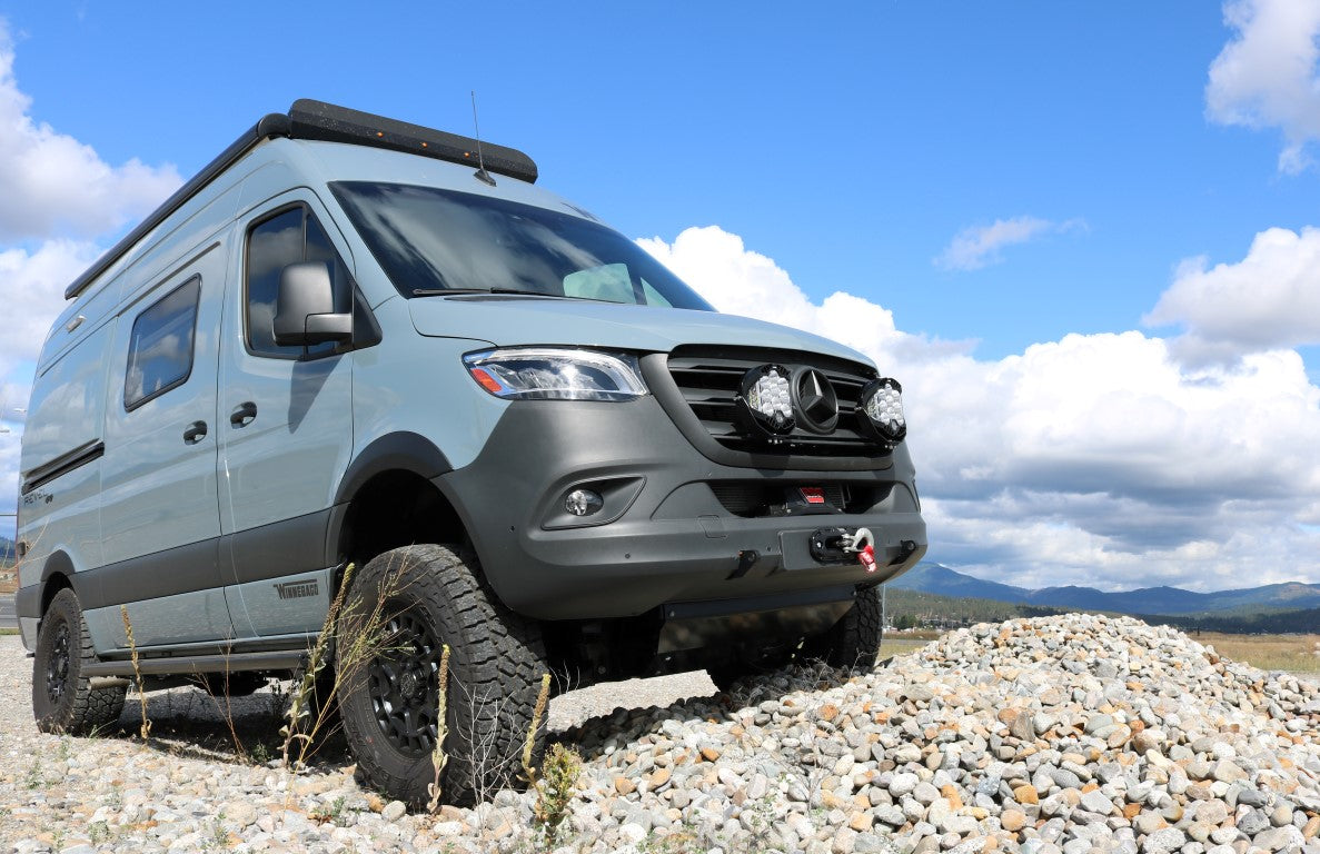 Floating Light Mount for 2019-present Sprinter by Van Compass on a grey van, positioned on rocks, showcasing a sleek, aggressive front-end lighting setup.
