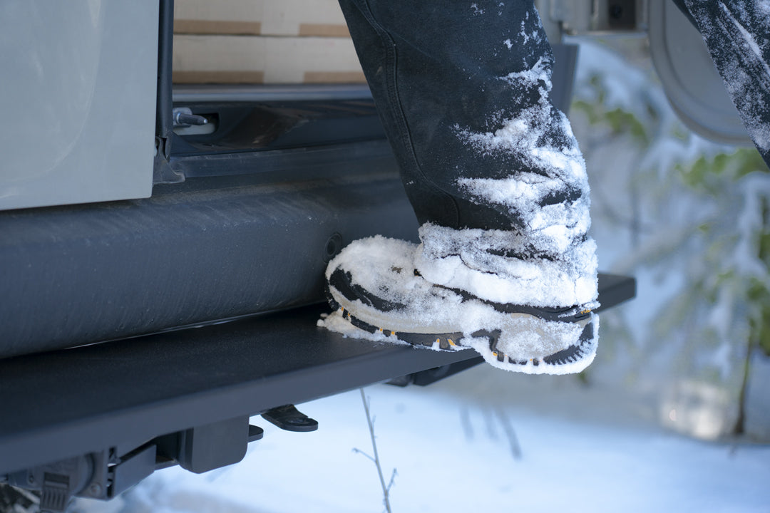 Close-up of a person's foot in snow, highlighting the REAR TUG STEP - SPRINTER (2019+) by VAN COMPASS attached to a vehicle's rear hitch.