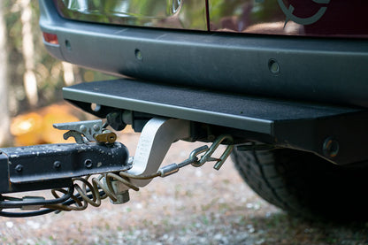 REAR TUG STEP - SPRINTER (2019+) by VAN COMPASS: Close-up of a metal tow step attached to a car's rear hitch, designed for easy cargo access and added vehicle protection.