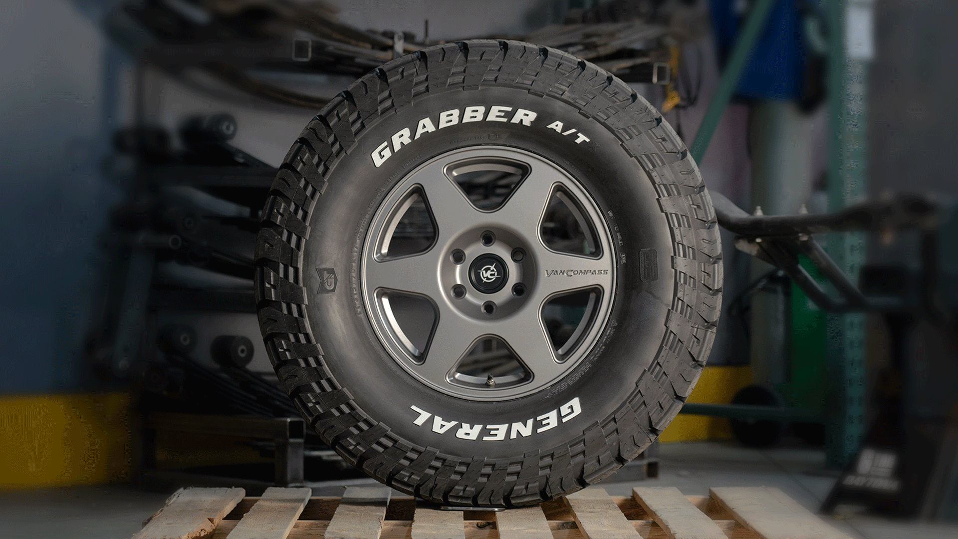 Animated rotating view of the Van Compass SLICE 17” alloy wheel mounted with a General Grabber A/T tire, featuring a sleek, modern five-spoke design and sharp detailing, shown in a workshop setting.