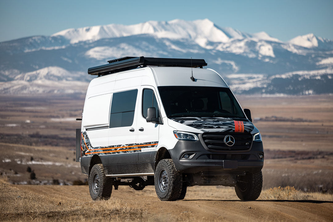 White van with black stripes on a dirt road, showcasing the Stage 5 System, 2 Lift - Sprinter AWD by Van Compass. Snowy mountains in the background.