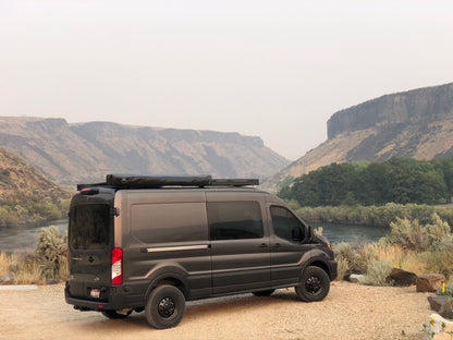 STAGE 4 TOPO 2.0 SYSTEM - TRANSIT RWD (2013+ SINGLE REAR WHEEL) by VAN COMPASS: Black van with enhanced suspension, parked on rocky terrain, showcasing improved ride quality and handling.