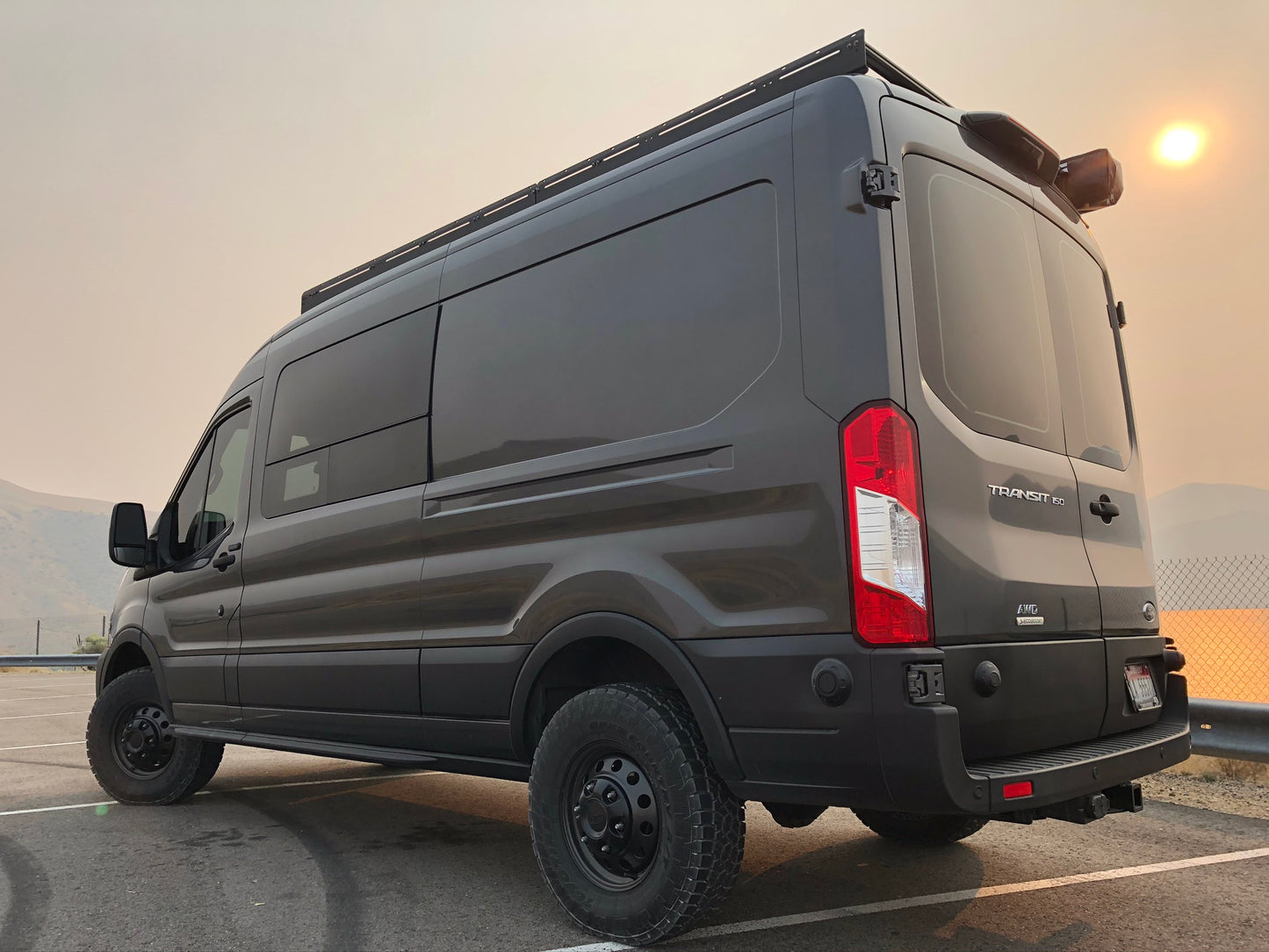 Black van equipped with the STAGE 3 TOPO 2.0 SYSTEM - TRANSIT RWD (2013+ SINGLE REAR WHEEL) by VAN COMPASS, parked in a lot, highlighting its enhanced stance and tire setup.