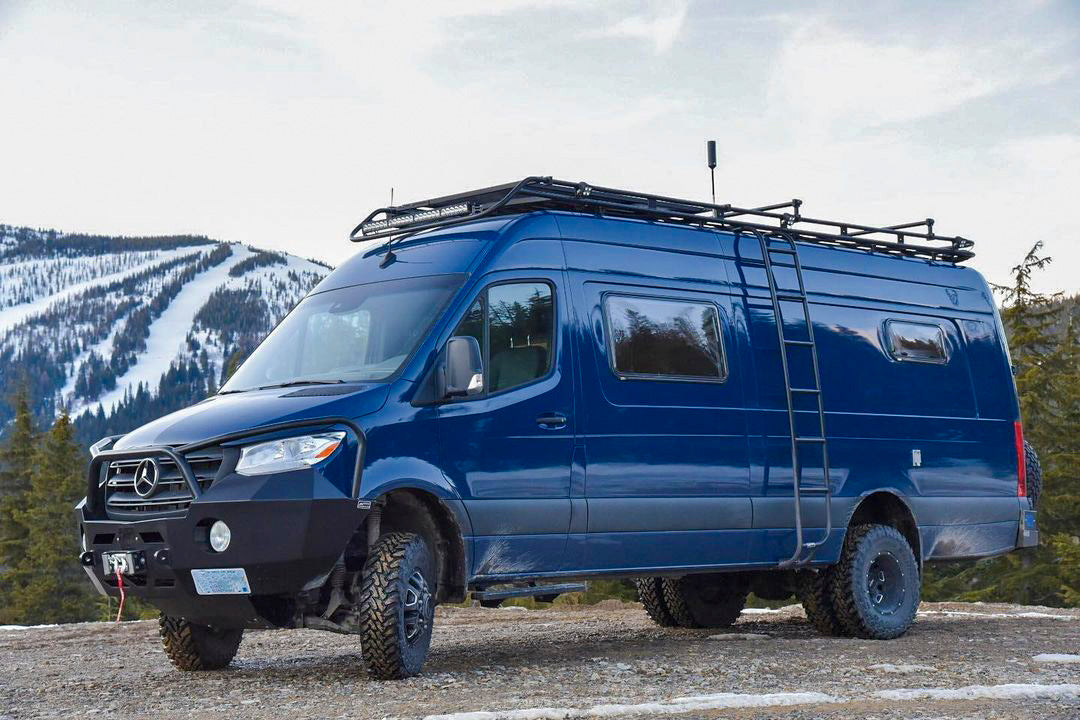 STAGE 6.3 DUALLY 2 LIFT SYSTEM - SPRINTER 4X4 (2019-2022 3500) by VAN COMPASS: Blue van on rocky road, showcasing high clearance and sturdy tires.